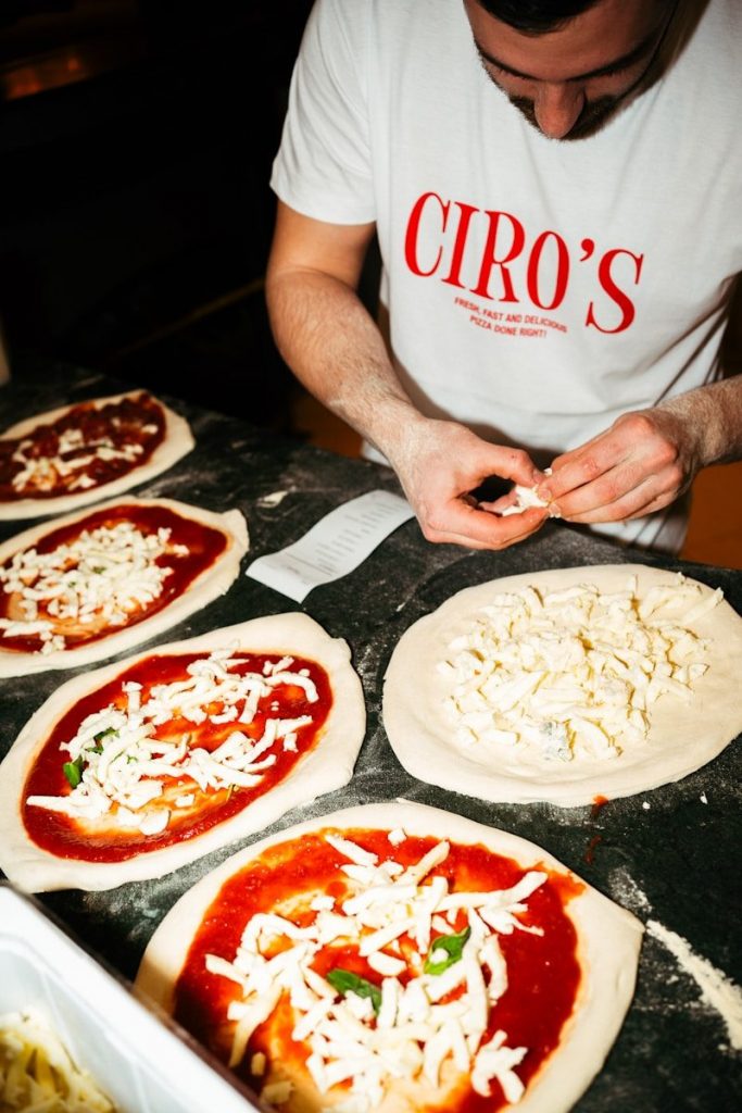 A person in a white t-shirt adding cheese to pizza dough on a counter.
