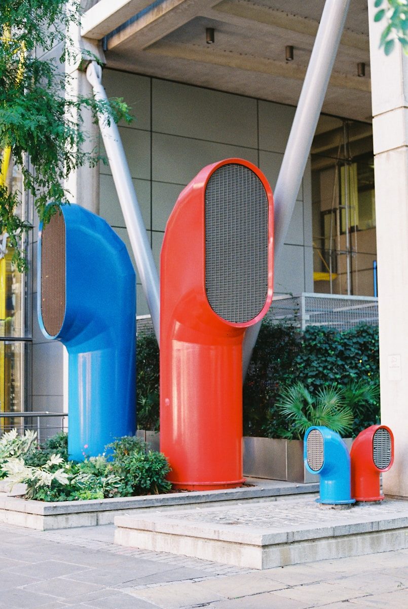 Large blue and red ventilation ducts standing among green plants outside a building